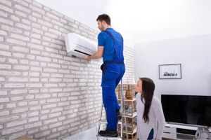 Professional technician repairing an air conditioning unit on a wall, with a woman observing, illustrating the importance of hiring specialists for HVAC services.