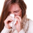 Person experiencing allergy symptoms while blowing nose into tissue with pained expression.