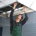 Male HVAC technician in green overalls inspecting ceiling tiles for leaks at Atlantic Heating & Air Conditioning.