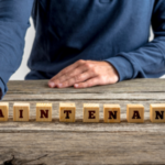 A person's hand arranging wooden letter blocks spelling "MAINTENANCE" on a wooden table, highlighting HVAC care.