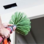 Person using a green duster for cleaning, with a ladder in background, promoting HVAC spring maintenance.