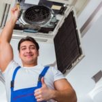 Smiling male technician in blue overalls gives thumbs up next to an indoor air conditioning unit during maintenance.