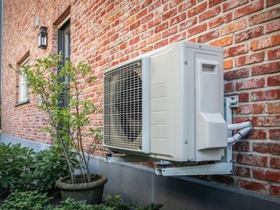 Outdoor air source heat pump mounted on a brick wall, surrounded by greenery, illustrating energy-efficient heating and cooling solutions for homes.