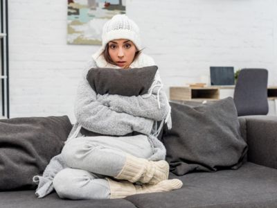 Woman sitting on a couch, bundled in warm clothing, holding a pillow, looking uncomfortable in a cold indoor setting, reflecting issues of uneven heating and humidity in homes.