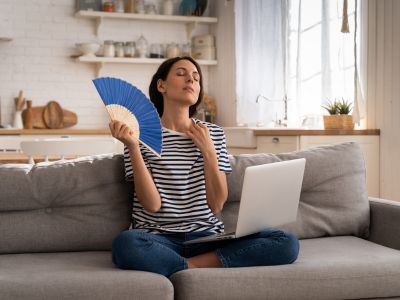 Woman sitting on a couch in a modern kitchen, using a blue fan for cooling while working on a laptop, illustrating the importance of effective refrigerant in home cooling systems.