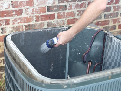 Person cleaning air conditioning coils with a spray nozzle, demonstrating maintenance to improve cooling efficiency and reduce energy costs.
