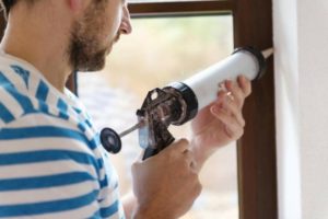 Man applying caulk to a window, demonstrating home maintenance for energy conservation and heat retention during winter.