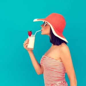 Woman in a striped swimsuit and wide-brimmed hat sipping a smoothie with a strawberry, against a vibrant turquoise background, evoking summer relaxation and comfort.