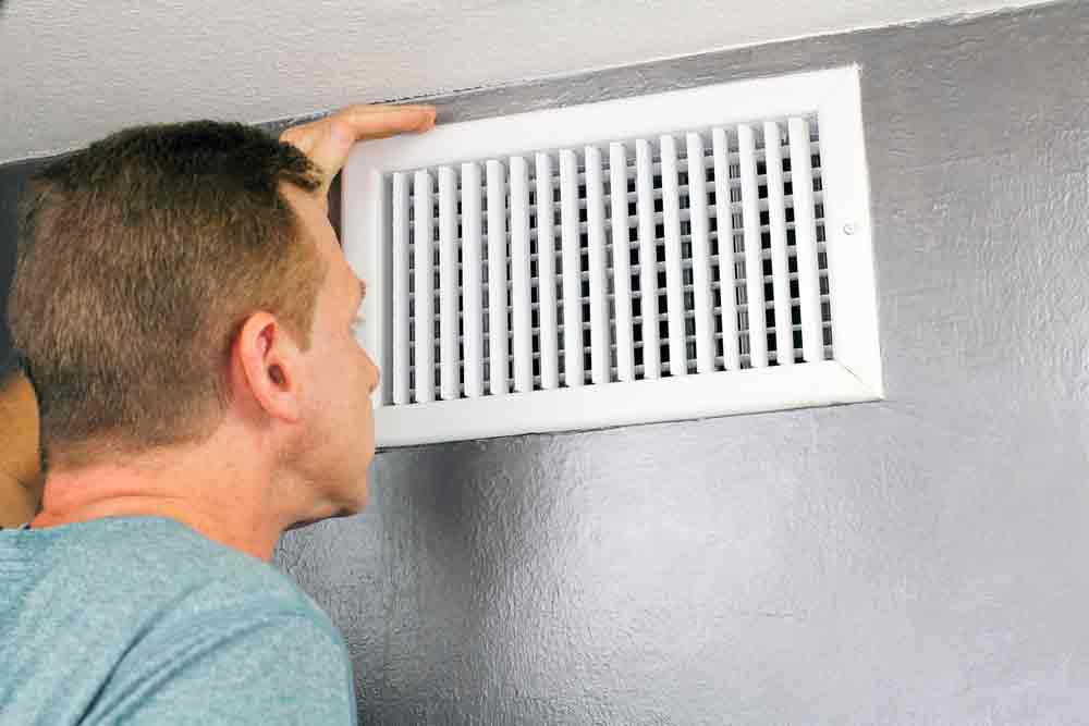 Man inspecting a wall vent for airflow issues in a home, addressing heating problems related to HVAC systems.